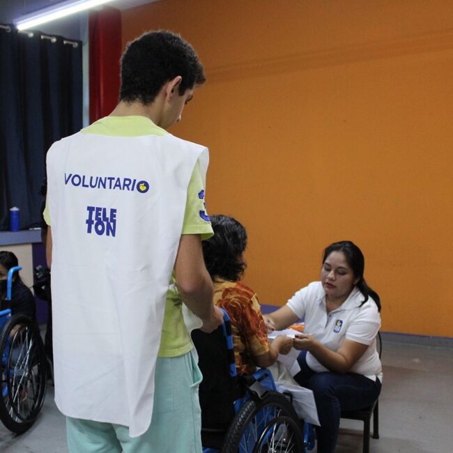 Voluntario de espalda ayudando a usuaria y terapeuta en entrega de donación de silla de ruedas.