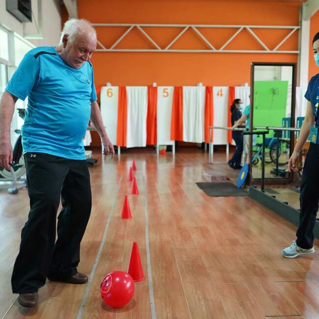 Hombre adulto mayor recibiendo terapia física a punto de patear un balón.